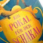 Text: "Ein Pokal für den Schakal". Eine Illustration eines Schakals, der auf einem gelben Pokal sitzt.