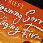Text: "Drowning Sorrows in Raging Fire", Illustration zeigt stilisierte Szene in warmen Farben mit einer Figur im Profil.