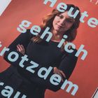 Text: "Heute geh ich trotzdem raus." Eine Frau mit verschränkten Armen vor einem orangefarbenen Hintergrund.