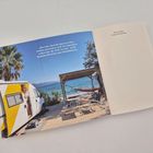 „Ein Auto, dem die Spuren seines Lebens anzusehen sind...“ Gelber Camper, Palme, Meer, Hängematte und mehrere Stühle.