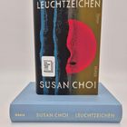 Buchcover: "LEUCHTZEICHEN", "SUSAN CHOI", "Piper". Unter dem Buch in Blau dieselben Texte. Kreis und Streifenmuster.
