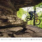 Kalender zeigt den Mai. Ein Radfahrer springt mit seinem Fahrrad über eine Felsformation im Wald.