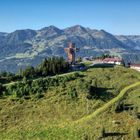 Ich kann keine Texte im Bild erkennen. Eine grüne Berglandschaft mit einem großen Holzkreuz und einigen Gebäuden vor einer Bergkette.
