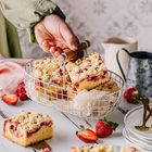 Keine sichtbaren Texte. Es sind Streuselkuchenstücke in einem Drahtkorb, umgeben von frischen Erdbeeren und Geschirr.