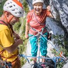 Zwei Personen klettern an einer Felswand. Sie tragen Schutzhelme und Gurte, die mit Seilen gesichert sind.