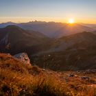 Sonnenuntergang über einer Berglandschaft, mit orange und violett gefärbtem Himmel und Licht auf den Berghängen.