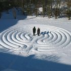 Zwei Personen stehen in einem schneebedeckten Labyrinth. Bäume umgeben das Labyrinth im Hintergrund.