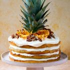 Schichttorte mit weißer Creme, dekoriert mit Nüssen und Ananas, belegt mit einer Ananas-Krone auf einem weißen Teller.