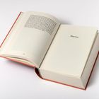 Der Text zeigt "Part two". Ein geöffnetes Buch mit rotem Einband liegt auf einer weißen Fläche.