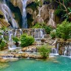 Ein mehrstufiger Wasserfall fließt über Felsen in klares, türkisfarbenes Wasser; umgeben von üppigem, grünem Dschungel.