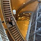 Eine Frau steigt eine breite, spiralförmige Treppe aus Metall und Holz in einem Gebäude mit orangefarbenen Wänden hinauf.