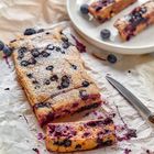 Blaubeerkuchen auf Backpapier, mit einer Scheibe abgeschnitten. Ein paar Blaubeeren verstreut, ein Messer daneben.