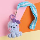 Ein gehäkelter Oktopus-Schlüsselanhänger mit lila Schleife lächelt fröhlich. Der Hintergrund ist bunt und geometrisch.