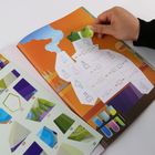 Zahlen und mathematische Symbole sind auf einer bunten Buchseite zu sehen. Eine Hand bewegt ein kleines, grünes Papierstück.