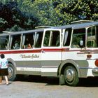 Der Text "Wanderfalke" steht auf einem Bus. Personen steigen ein. Bäume im Hintergrund. Retro-Stil.