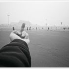 Eine ausgestreckte Hand zeigt den Mittelfinger auf einen großen, menschengefüllten Platz in einer nebligen Umgebung.