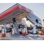 Text: "AVIA" auf einem überlasteten Tankstellenvordach mit mehreren Fahrzeugen. Eine Säule ist schief und trägt Autos.