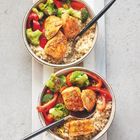 Zwei Schalen mit Quinoa, gebratenem Huhn, roten Paprikastreifen und Brokkoli. Ein schwarzer Löffel liegt in jeder Schale.