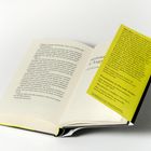 Gelber Zettel mit Text über den Autor Alex Beer, vor einem aufgeschlagenen Buch platziert.