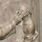 Eine marmorne Skulptur zeigt eine Person, die auf eine andere weist, die ihre Augen berührt. Beide in fließenden Gewändern.