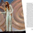 Ein Performer mit langem Haar, in einem funkelnden weißen Anzug, singt in ein Mikrofon vor großen, spiralförmigen Dekorationen.