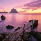 Ein hölzerner Steg führt von felsigen Uferabschnitten ins ruhige, pink-violette Meer, dahinter ein großer Felsen im Sonnenuntergang.