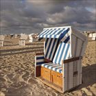 Strand mit mehreren Strandkörben, vorderer Korb blau-weiß gestreift. Wolkiger Himmel im Hintergrund.