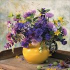 Keine Texte vorhanden. Ein gelber Krug voller bunter Blumen, hauptsächlich in Lila- und Gelbtönen, auf einem Holztablett.