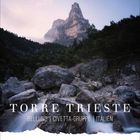 "Torre Trieste" mit Berg im Hintergrund, bewaldete Hügel und Steine im Vordergrund, ruhiger Wasserlauf.