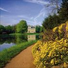 Pfad mit gelben Blumen in einem Park; im Hintergrund ist ein größeres Gebäude am Ufer eines Sees zu sehen.