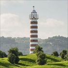 Leuchtturm mit rot-weißen Streifen auf grünem Hügel, bewachsen mit kleinen Bäumen, im Hintergrund sanfte Hügel.