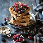 Ein Stapel Waffeln mit Blaubeeren, Himbeeren und Mandeln auf einem Teller. Eine Serviette und Schalen mit Toppings daneben.