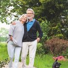 Ein älteres Paar spaziert Arm in Arm einen Gartenweg entlang. Ein kleiner Hund begleitet sie.