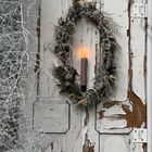 Eine Kerze brennt in einem Kranz an einer alten, schäbig-weißen Tür. Daneben sind vereiste Zweige sichtbar.