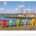 Lunenburg, Nova Scotia. Fünf bunte Stühle auf einem Holzsteg vor Wasser und Segelschiffen. Darunter ein Kalender für Mai.