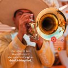 „Das Leben ist kein Wunschkonzert, aber manchmal spielt es dein Lieblingslied.“ Ein Mann spielt Trompete, bunte Blumen.