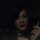 Eine Frau im Dunkeln, trägt roten Lippenstift und Pelzmantel, hält eine Zigarette. Ihr Gesichtsausdruck wirkt nachdenklich.