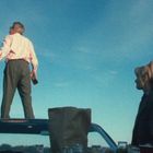 Keine erkennbaren Texte. Ein Mann steht auf einem Autodach, hält eine Flasche, eine Frau mit Pferdeschwanz schaut zu.