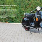 „Erste Liebe rostet nicht“; Diane besitzt ihre P 80 X seit sie sechzehn ist. Schwarze Vespa auf gepflastertem Boden vor Hecke.
