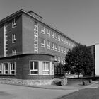 Text: "Fabricius". Ein mehrstöckiges, rechteckiges Backsteingebäude mit vielen Fenstern, Bäume und eine gepflasterte Straße.