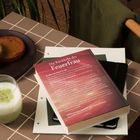 Der Text auf einem Buch lautet: "Die Rückkehr der Feuerfrau." Daneben ein Getränk und zwei Kekse auf einem Tisch.