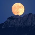 Ein großes, leuchtend gelbes Vollmond steht hinter einer felsigen Bergspitze mit einem Kreuz darauf.