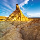 Kein Text vorhanden. Ein markanter Felsen in einer Wüstenlandschaft unter einem strahlend blauen Himmel mit zarten Wolken.