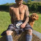 „Mai“ steht unten. Ein muskulöser Mann im Kilt auf Heuballen, neben ihm ein brauner Hund, grüne Wiese im Hintergrund.