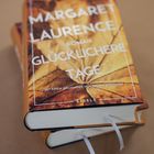 "Buchcover 'Margaret Laurence Roman Glücklichere Tage'. Darauf ein herbstliches Laubblatt, darunter zwei Bücher gestapelt."