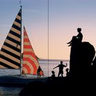 Segelboot mit gemusterten Segeln auf Wasser bei Sonnenuntergang, Silhouetten von Menschen auf Felsen im Vordergrund.