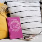 Das Buch "FREUDE: Das Leben ins Herz schließen" von Melanie Wolfers in Pink liegt auf einem gelben Kissen vor einem gestreiften.