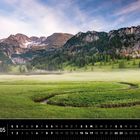 Kalender mit Landschaftsfoto der Alpen im Mai. Grüne Wiesen, schlängelnder Bach, Berge im Hintergrund, blauer Himmel.