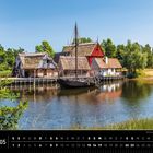 Dänemark 05: Historische Gebäude mit Reetdächern an einem See, traditionelles Holzboot im Wasser, umgeben von grüner Natur.