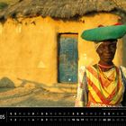 „Traditionell gekleidete Herero-Frau in Warmquelle, Kaokoveld“. Eine Frau in bunter Kleidung vor einer Hütte. Unten: „Namibia 05“.